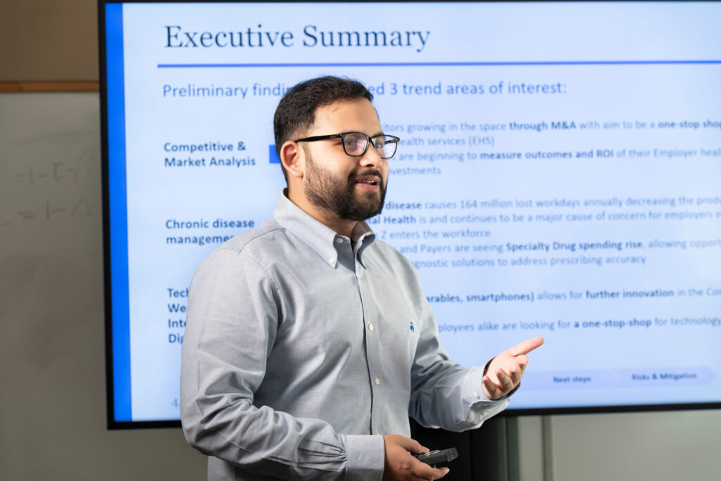 A student gestures while he presents information in front of a screen that says Executive Summary